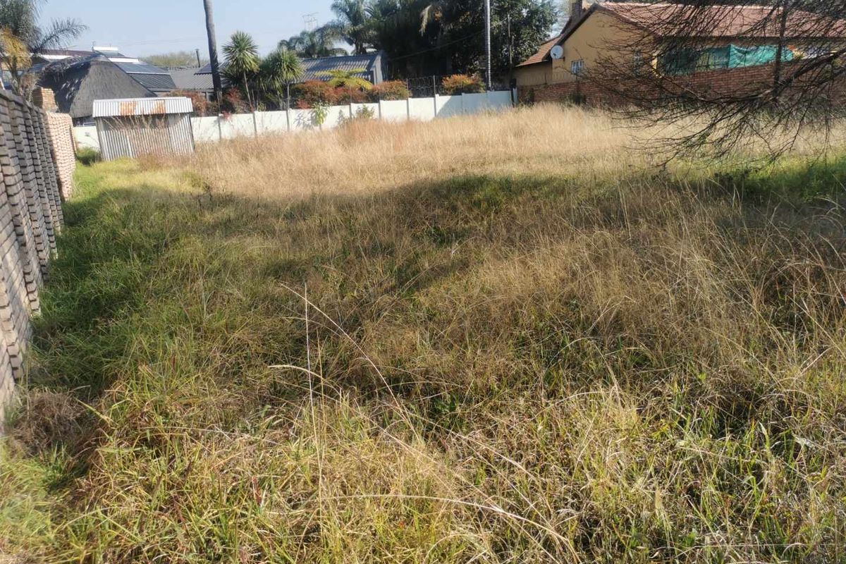 A vacant residential plot featuring overgrown grass, enclosed by perimeter brick walling and fencing in a suburban neighborhood setting.