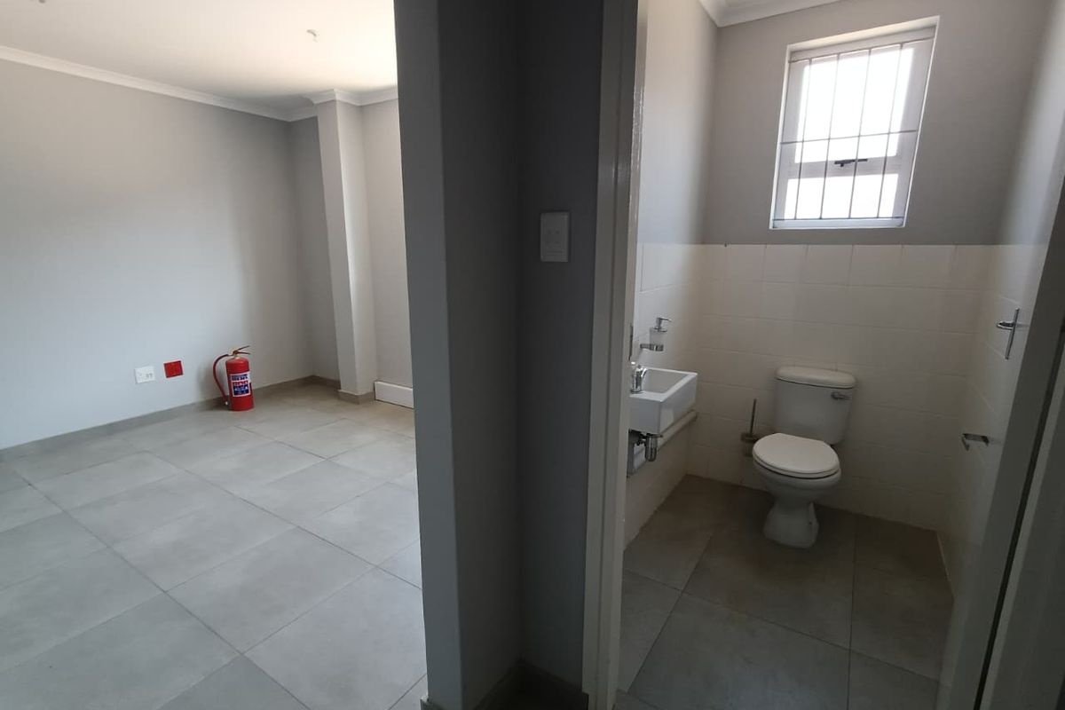 A clean, functional bathroom featuring a wall-mounted basin and toilet, adjacent to an open-plan office or utility space with tiled flooring.