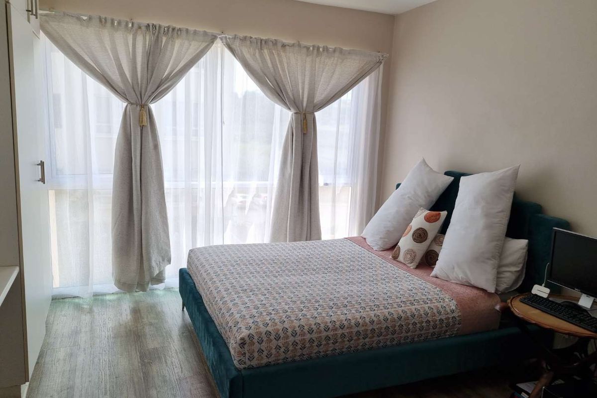 A well-lit bedroom featuring wood-look flooring, built-in wardrobe storage, and large windows with sheer curtains.