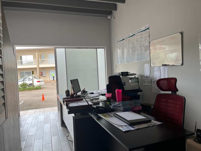 A functional office space featuring tiled flooring, exposed ceiling beams, and a sliding glass door providing direct access to the exterior parking area.