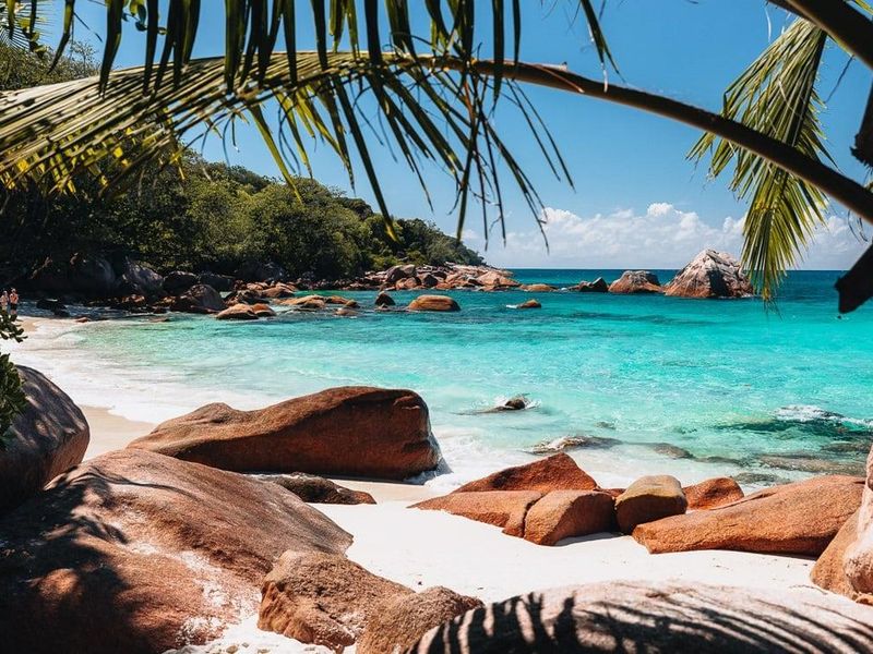 A scenic view of a pristine tropical beach with turquoise waters, white sand, and large granite boulders, framed by palm fronds.