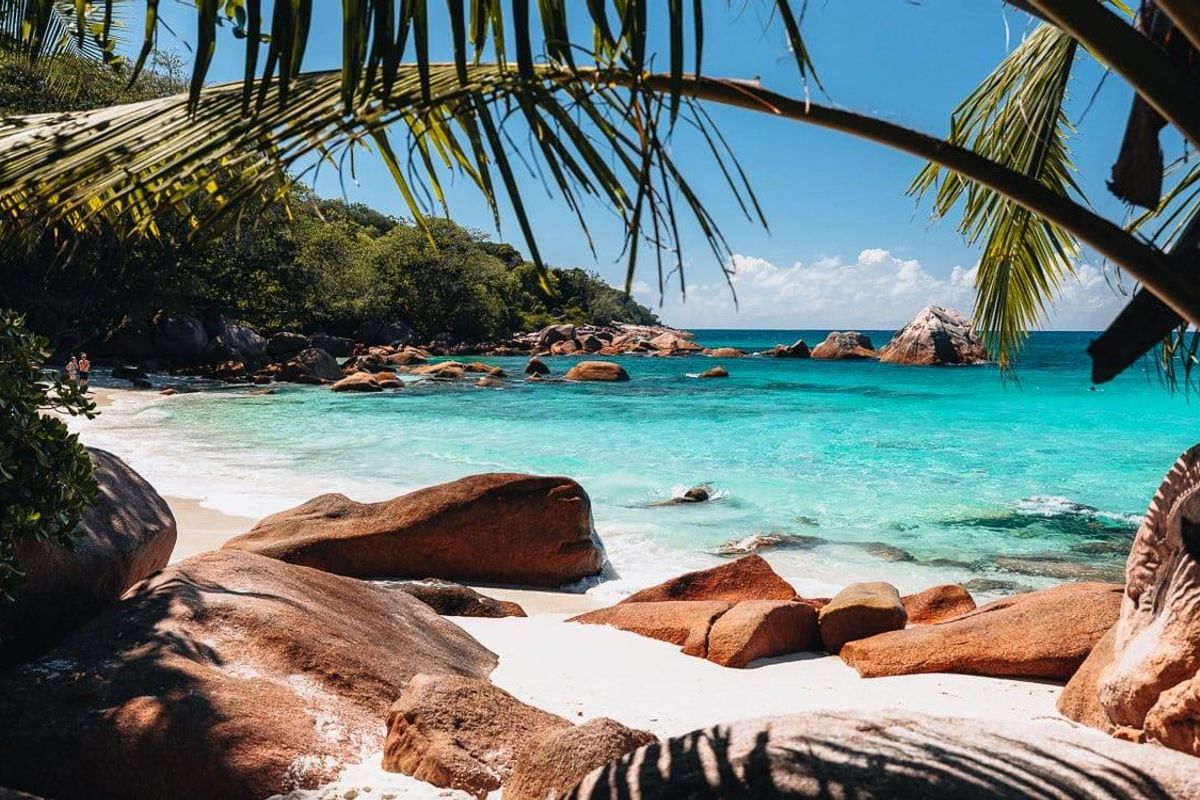 A scenic view of a pristine tropical beach with turquoise waters, white sand, and large granite boulders, framed by palm fronds.