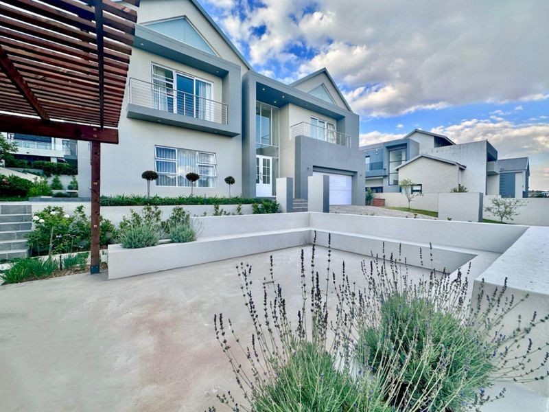 Modern double-storey residence featuring a spacious concrete patio with built-in seating, a timber pergola, and well-maintained landscaped garden beds.