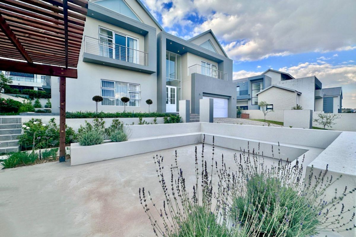 Modern double-storey residence featuring a spacious concrete patio with built-in seating, a timber pergola, and well-maintained landscaped garden beds.