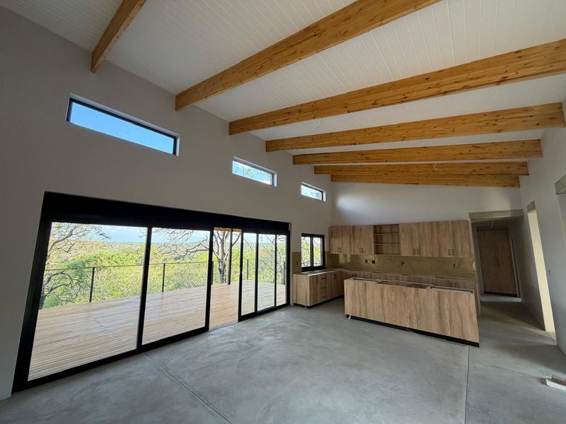 Spacious open-plan kitchen and living area featuring exposed timber beams, polished concrete floors, and large sliding doors opening onto a scenic timber deck.