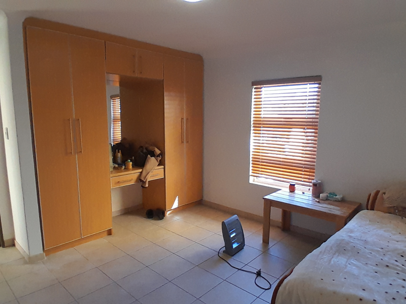 A bedroom featuring built-in wooden cupboards with an integrated vanity, tiled flooring, and a window fitted with venetian blinds.