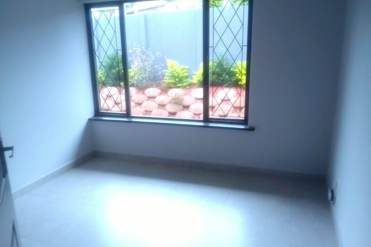 A bright, empty bedroom featuring tiled flooring and a large window with security bars overlooking a landscaped garden area.