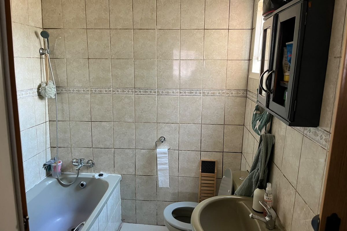 A compact bathroom featuring full-height wall tiling, a bathtub with shower mixer, a pedestal basin, and a toilet, offering potential for modern renovation.