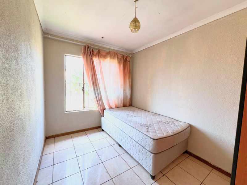 A compact bedroom featuring tiled flooring, a window with security bars, and a simple pendant light fixture, requiring cosmetic updates to improve its overall condition.
