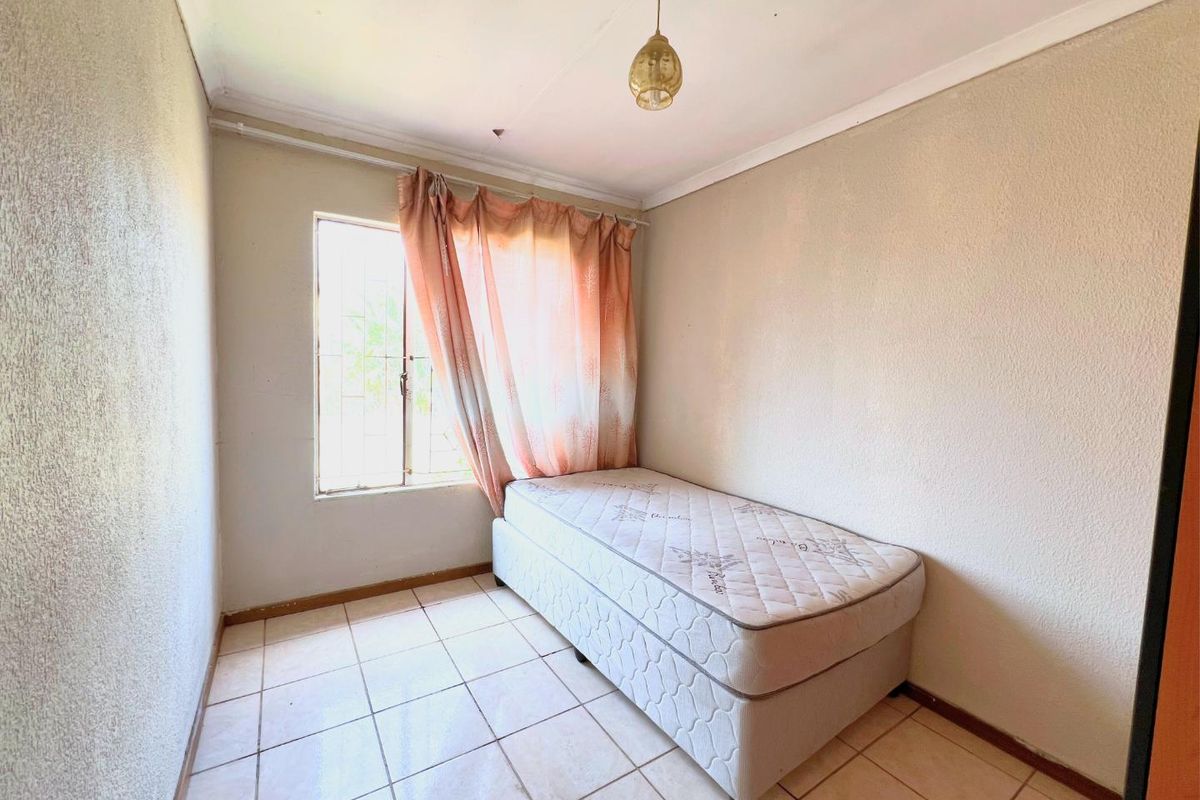 A compact bedroom featuring tiled flooring, a window with security bars, and a simple pendant light fixture, requiring cosmetic updates to improve its overall condition.