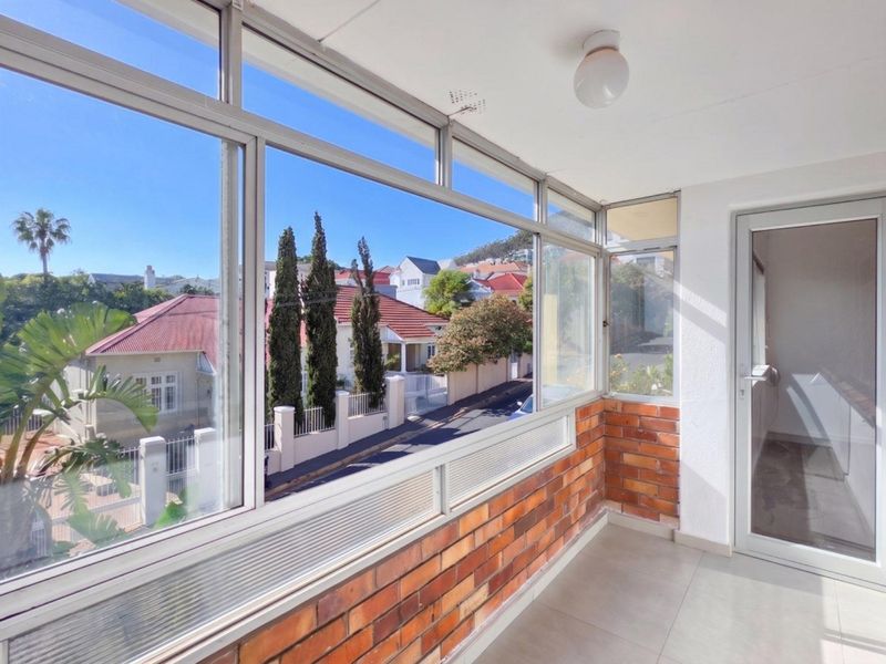 An enclosed balcony featuring tiled flooring, exposed brickwork, and large aluminium-framed windows offering a pleasant neighbourhood view.