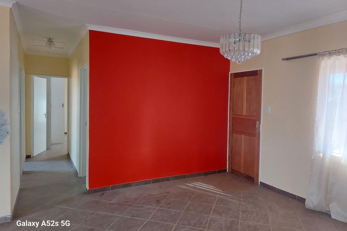 A living area featuring tiled flooring, a prominent red accent wall, and a central chandelier, with an adjacent hallway leading to other rooms.