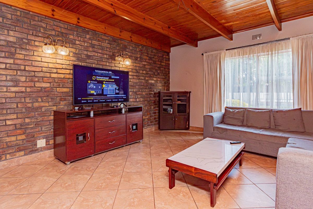 A living room featuring an exposed brick feature wall, timber beam ceiling, and tiled flooring with access to the outdoors via a sliding door.