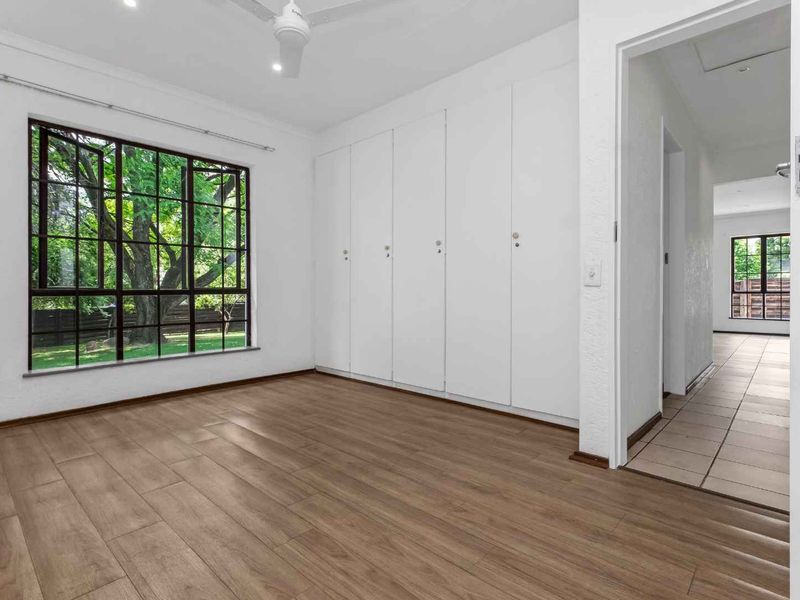 A bright, empty bedroom featuring built-in wardrobes, a ceiling fan, and modern laminate flooring, with a view into the adjacent tiled hallway.