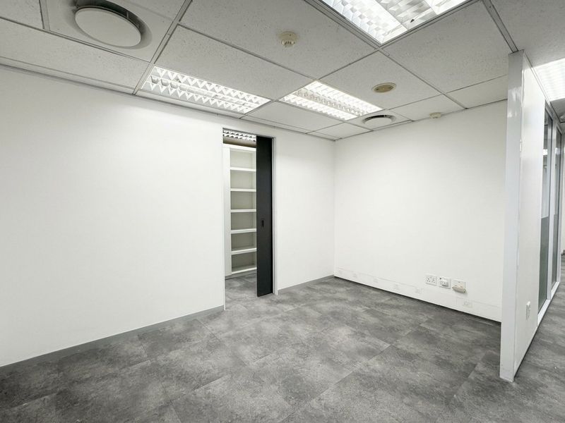 A clean, vacant office space featuring a suspended ceiling, recessed lighting, grey tiled flooring, and an adjoining storage area with built-in shelving.