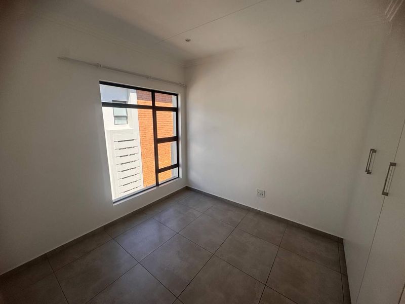 A bright, empty bedroom featuring neutral tiled flooring, built-in cupboards, and a large aluminium-framed window providing natural light.