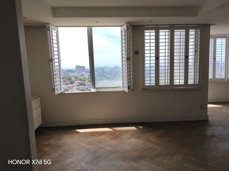 Bright and spacious living area featuring elegant herringbone timber flooring, plantation shutters, and expansive windows offering panoramic city views.