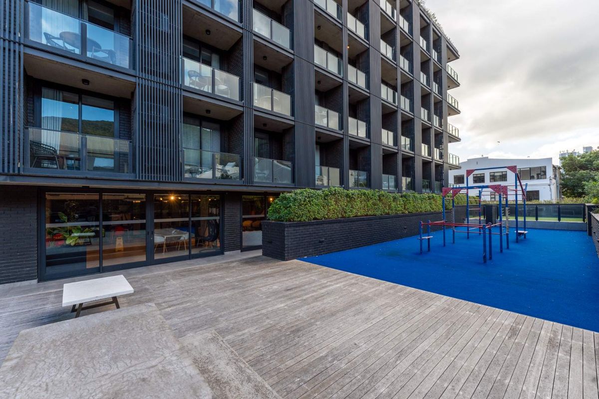 Rear exterior view of the apartment building featuring a timber deck, glass balcony railings, and a communal playground area with blue safety surfacing.