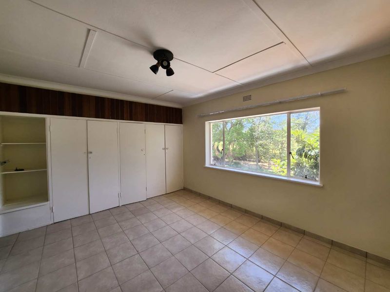 An empty bedroom featuring tiled flooring, built-in cupboards, and a large window overlooking the garden.