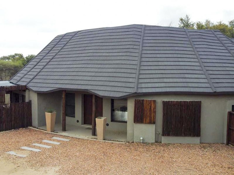 Front elevation of a house featuring a large tiled roof, covered entrance with timber posts, and a gravel driveway area.