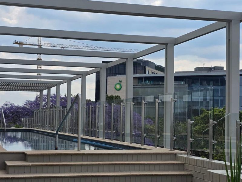A modern rooftop swimming pool area featuring a sleek glass balustrade, timber decking, and a structural metal pergola with urban views.