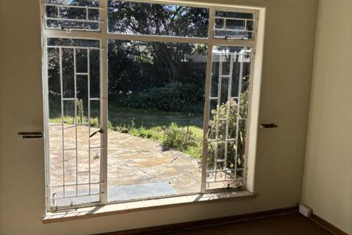 A bright, empty living room featuring large windows with security bars, offering a view of the rear patio and garden area.