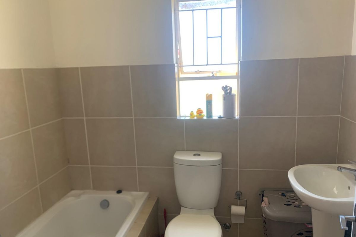 A functional bathroom featuring a built-in bathtub, pedestal sink, and toilet, finished with neutral wall tiling and a window with security bars.