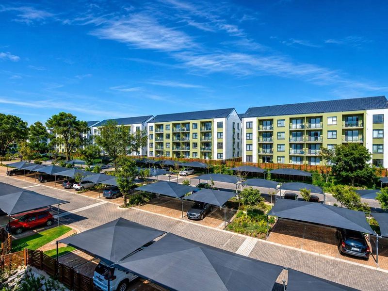 Aerial view of a modern apartment complex featuring multi-story residential buildings, paved parking areas with shade cloth carports, and landscaped grounds.