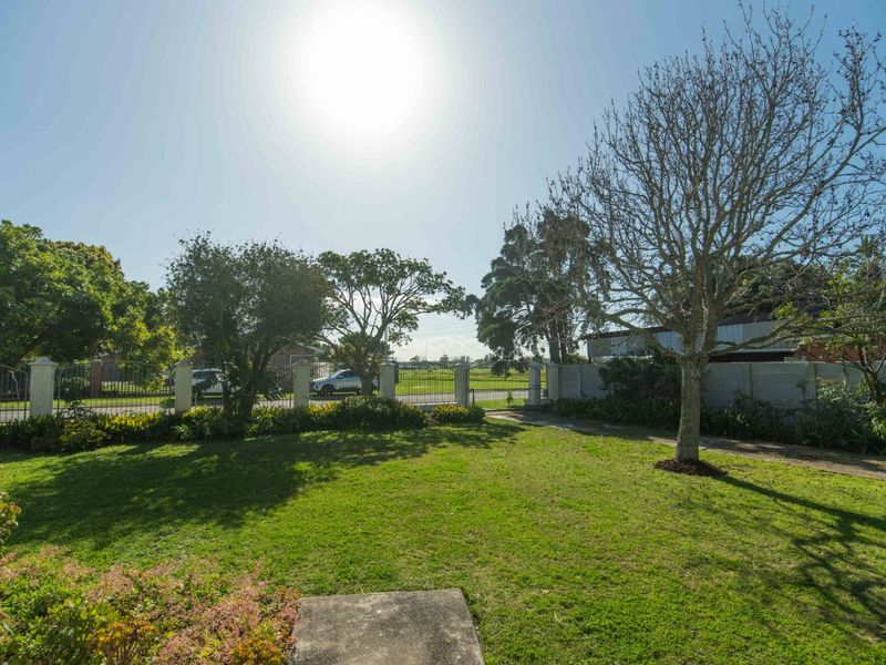 A spacious front garden featuring a well-maintained lawn, mature trees, and a secure perimeter wall with a metal entrance gate.