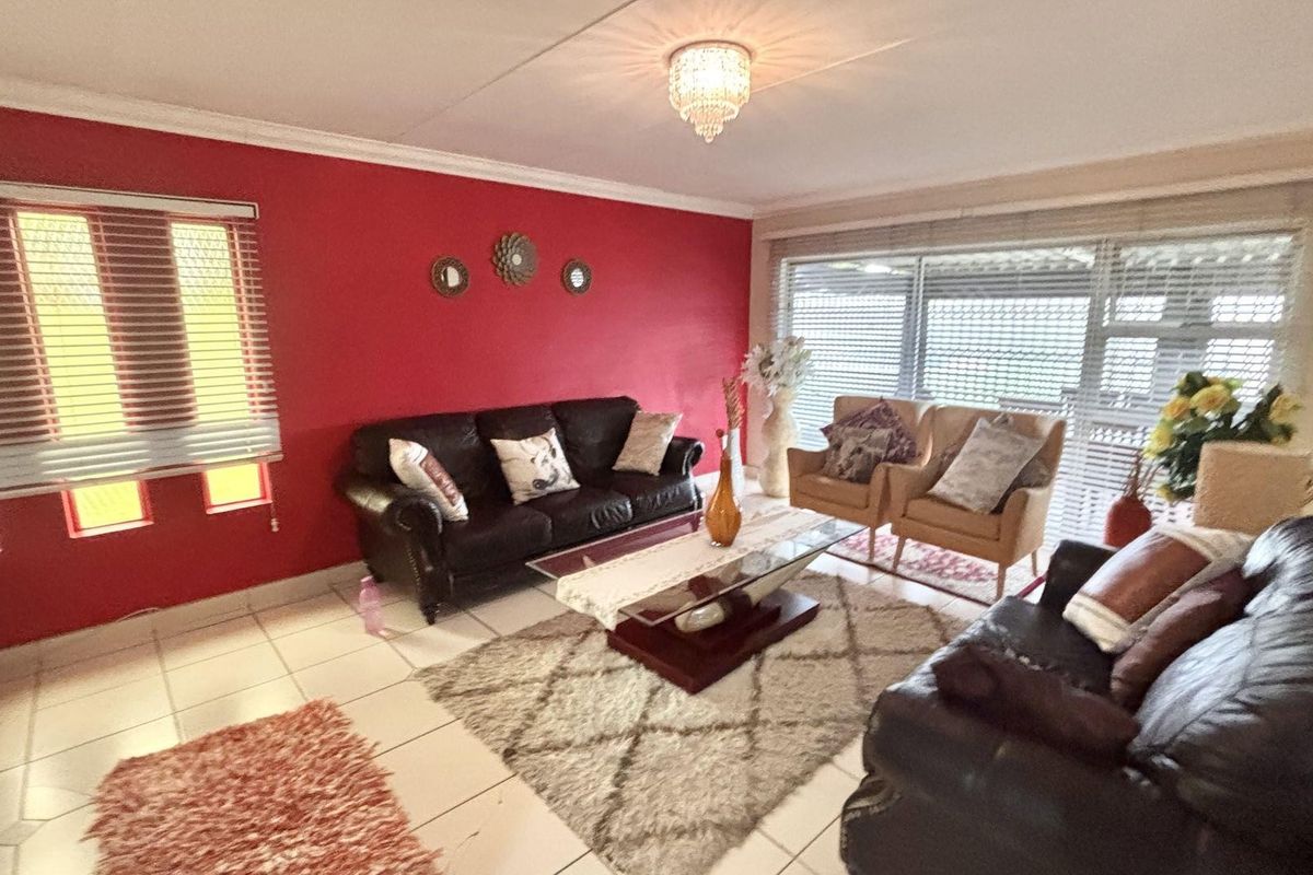 A spacious living room featuring tiled flooring, a bold red accent wall, and large windows with fitted blinds allowing for natural light.