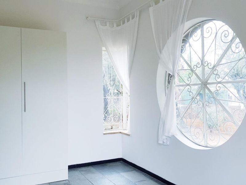 A bright, empty bedroom featuring a unique circular window with decorative security grille, built-in wardrobe, and tiled flooring.