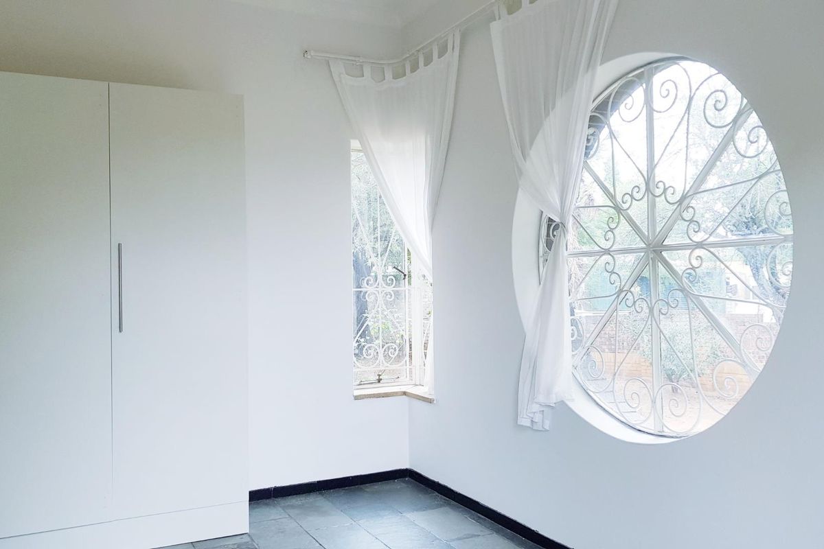 A bright, empty bedroom featuring a unique circular window with decorative security grille, built-in wardrobe, and tiled flooring.