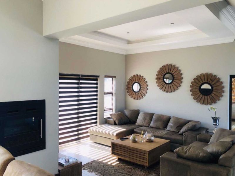 Spacious living room featuring a recessed tray ceiling, built-in fireplace, and modern tiled flooring with natural light filtering through venetian blinds.