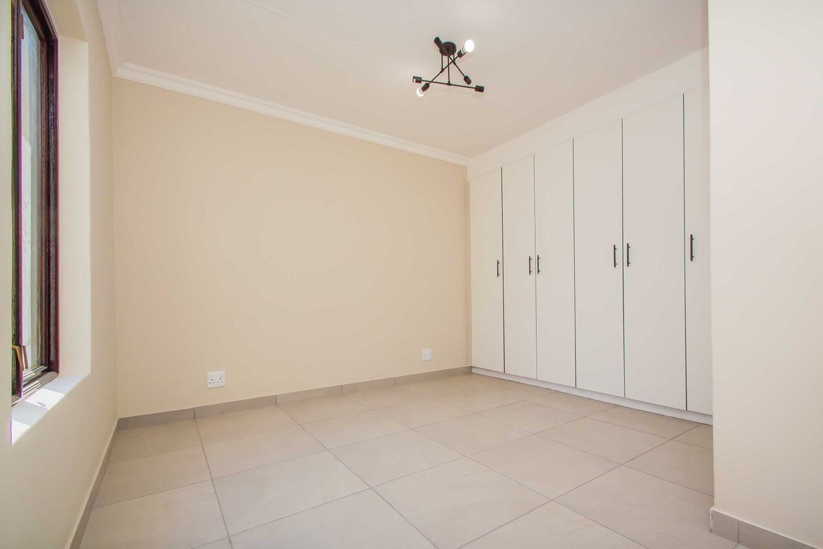 A bright, empty bedroom featuring neutral tiled flooring, built-in white cupboards, and a modern ceiling light fixture.