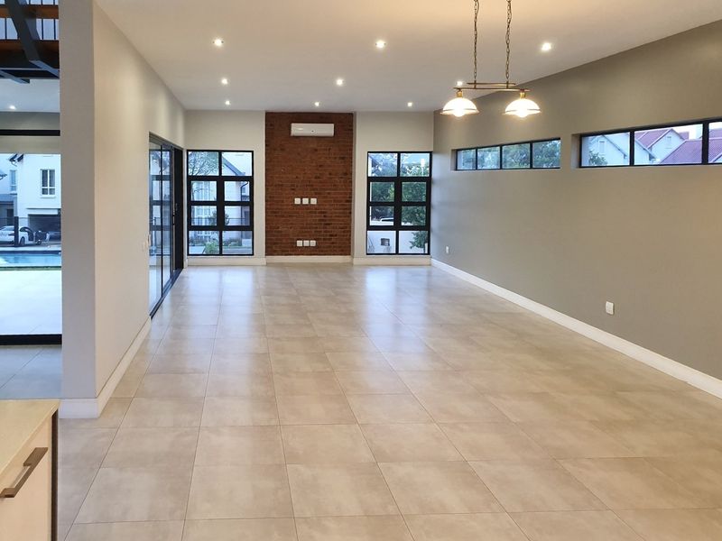 Spacious, modern open-plan living area featuring tiled floors, an exposed brick feature wall, recessed lighting, and large windows providing ample natural light.