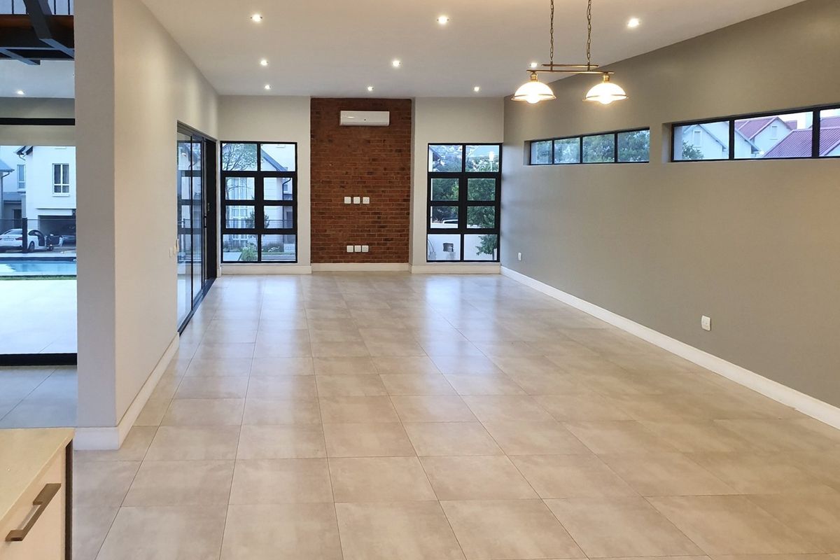 Spacious, modern open-plan living area featuring tiled floors, an exposed brick feature wall, recessed lighting, and large windows providing ample natural light.