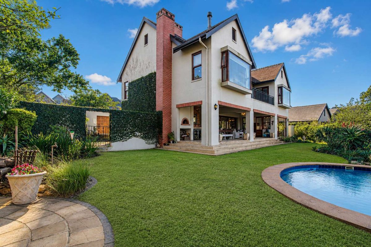Rear view of a multi-story house featuring a swimming pool, landscaped garden, covered patio with built-in braai, and a prominent brick chimney.