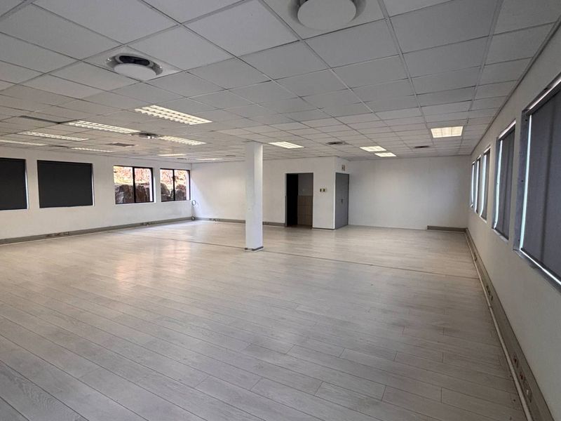 A spacious, open-plan office area featuring a suspended ceiling, recessed lighting, laminate flooring, and large windows with fitted roller blinds.