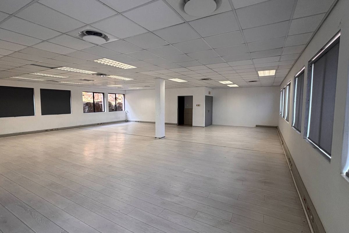 A spacious, open-plan office area featuring a suspended ceiling, recessed lighting, laminate flooring, and large windows with fitted roller blinds.