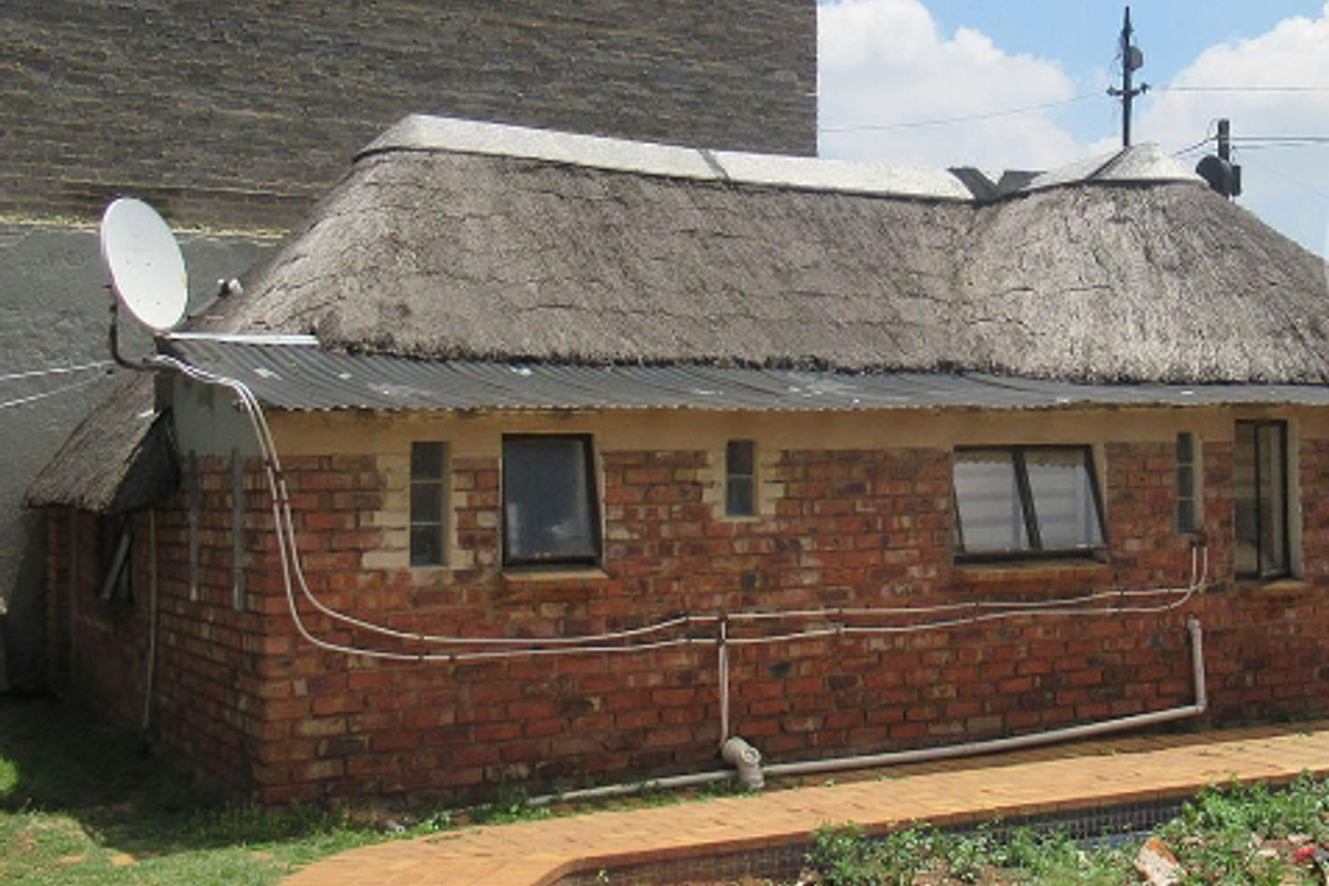 Exterior view of a brick-built structure with a traditional thatch roof, featuring external plumbing and wiring, adjacent to a paved walkway and garden.