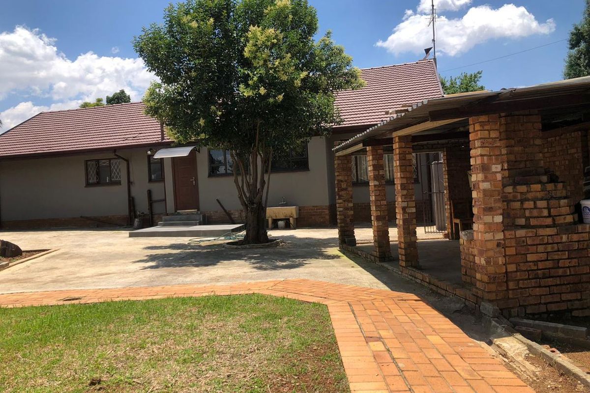 Rear exterior view featuring a paved courtyard, a covered brick patio area, and a well-maintained garden space with mature trees.