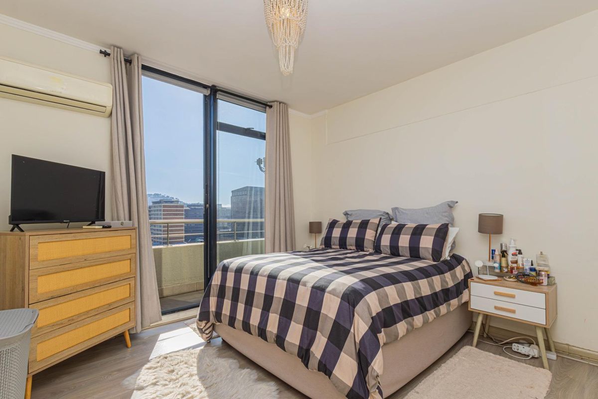 A bright bedroom featuring wood-look flooring, a wall-mounted air conditioning unit, and large sliding glass doors providing access to a private balcony with city views.