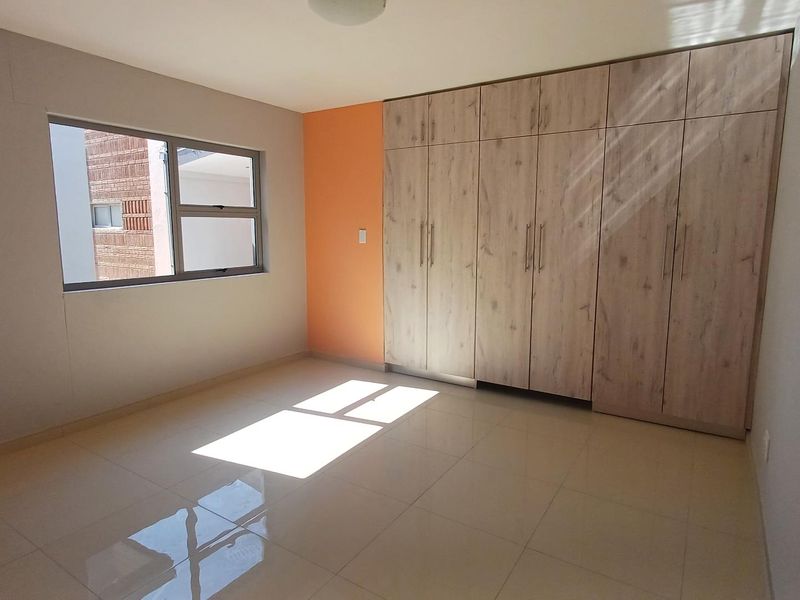 A bright, empty bedroom featuring modern light wood-finish built-in cupboards, neutral tiled flooring, and a large window allowing for ample natural light.