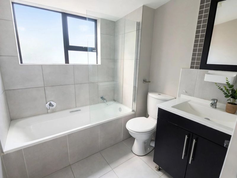 A modern, well-maintained bathroom featuring a bathtub with a glass screen, a vanity unit with a basin, and neutral floor-to-ceiling tiling.