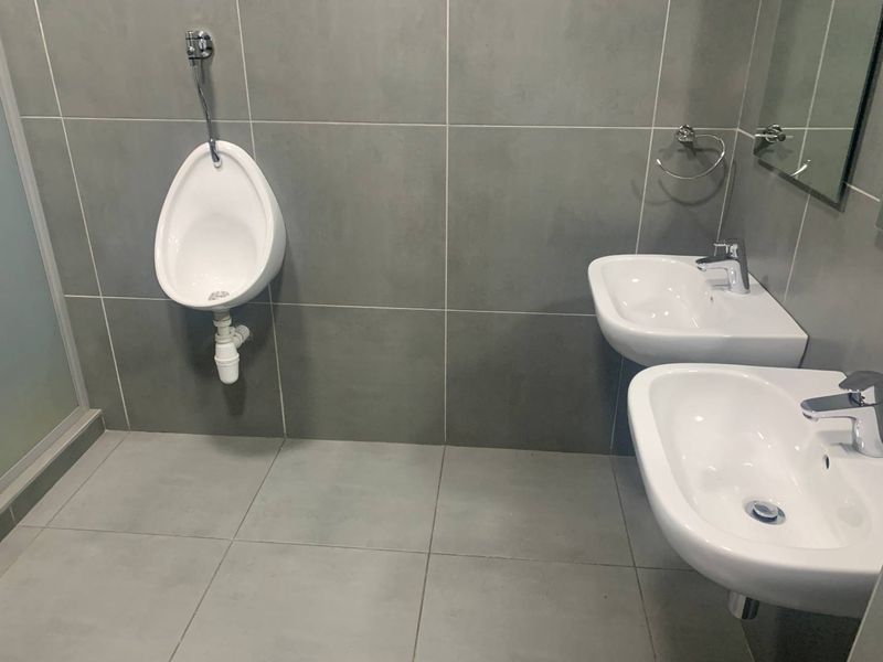 Modern office bathroom featuring dual wall-mounted washbasins with chrome fixtures and a wall-mounted urinal, finished with large format grey ceramic tiling.