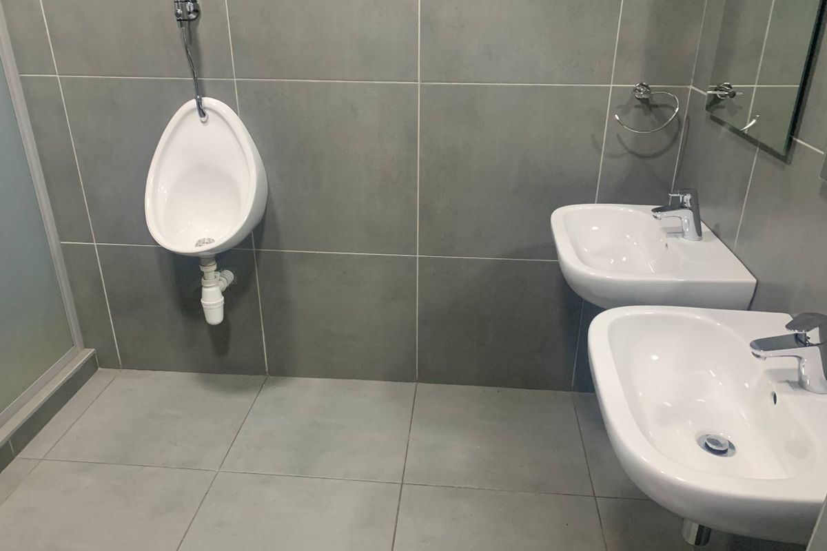 Modern office bathroom featuring dual wall-mounted washbasins with chrome fixtures and a wall-mounted urinal, finished with large format grey ceramic tiling.