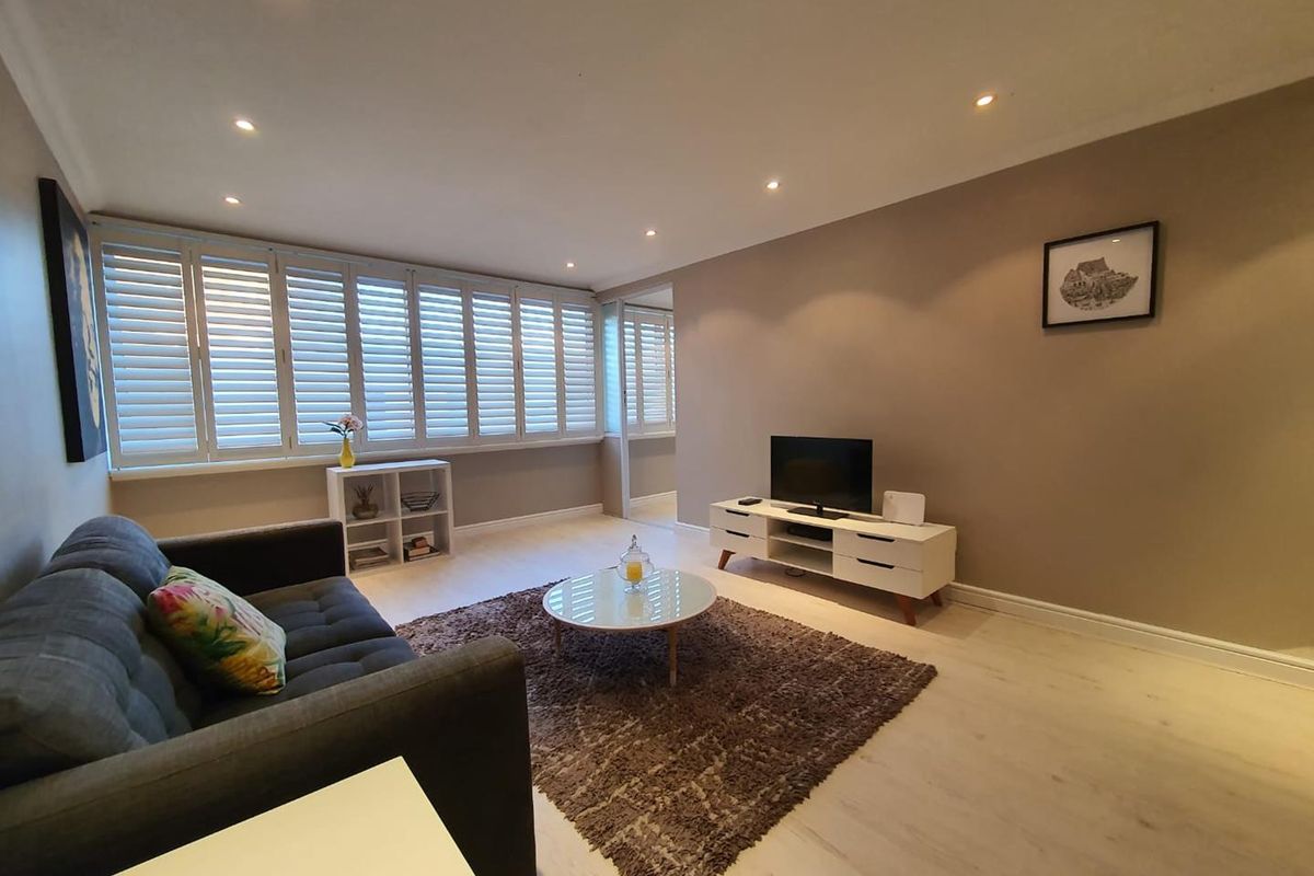 A bright and modern living room featuring light wood-look flooring, recessed ceiling lighting, and white plantation shutters on the windows.
