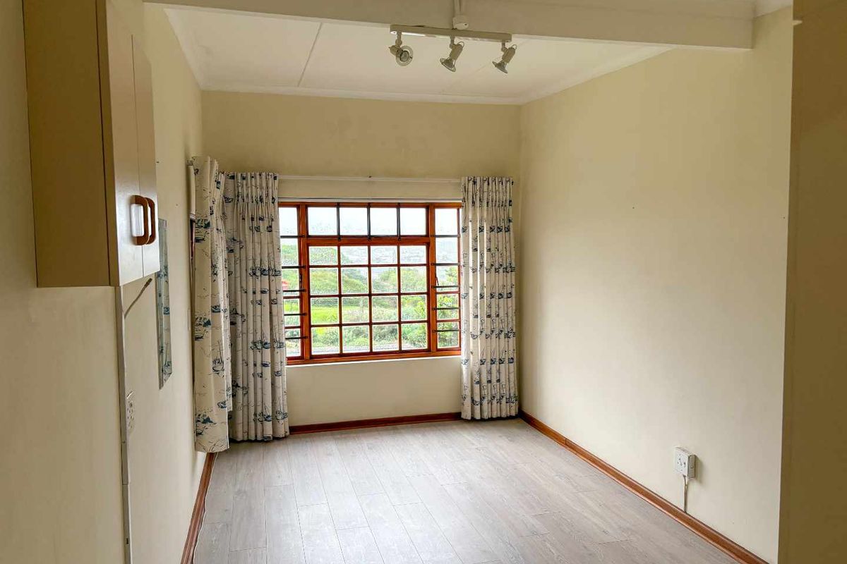 A bright, empty bedroom featuring light-toned laminate flooring, a wooden-framed window with security bars, and a built-in wall cabinet.