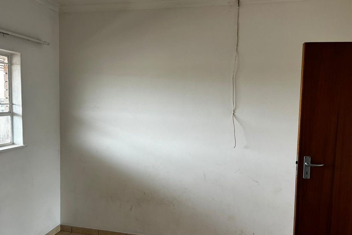A vacant bedroom featuring tiled flooring, a single window, and a wooden door, with visible wall scuffs and exposed electrical wiring.