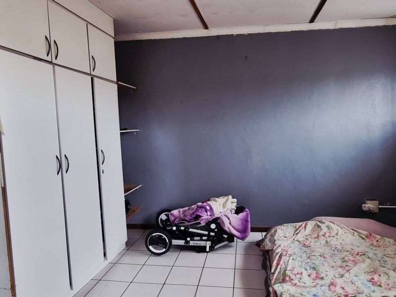 A bedroom featuring built-in cupboards, tiled flooring, and a window with security bars. The room requires cosmetic updates to the ceiling and walls.